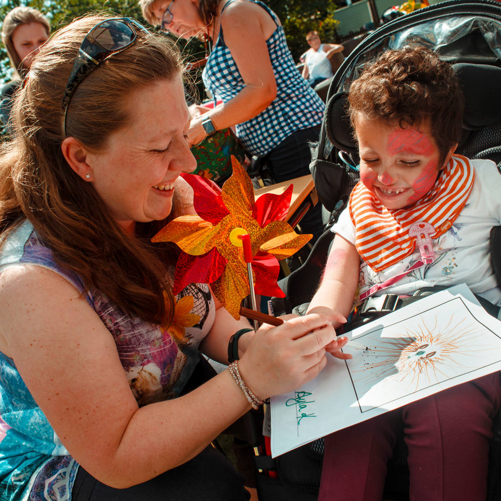 A smiling woman and a young child in a wheelchair enjoy an alphabet design activity together, surrounded by colorful decorations and other participants.
