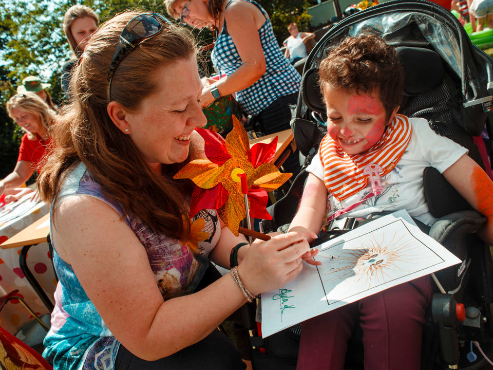 A smiling woman and a young child in a wheelchair enjoy an alphabet design activity together, surrounded by colorful decorations and other participants.
